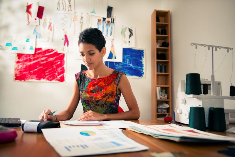 Money and financial planning, young hispanic self-employed woman checking bills and doing budget with calculator, computer and papers in fashion design studio