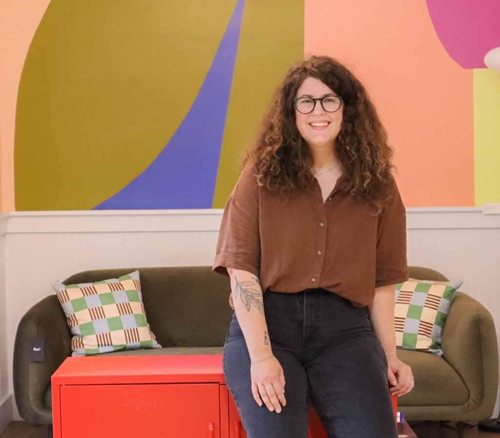 Sarah Lawrence poses in storefront. She is wearing a brown short with black pants and has curly brown hair with glasses.