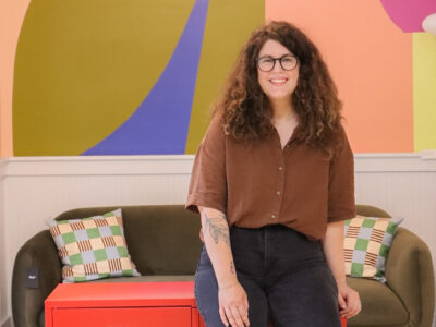 Sarah Lawrence poses in storefront. She is wearing a brown short with black pants and has curly brown hair with glasses.